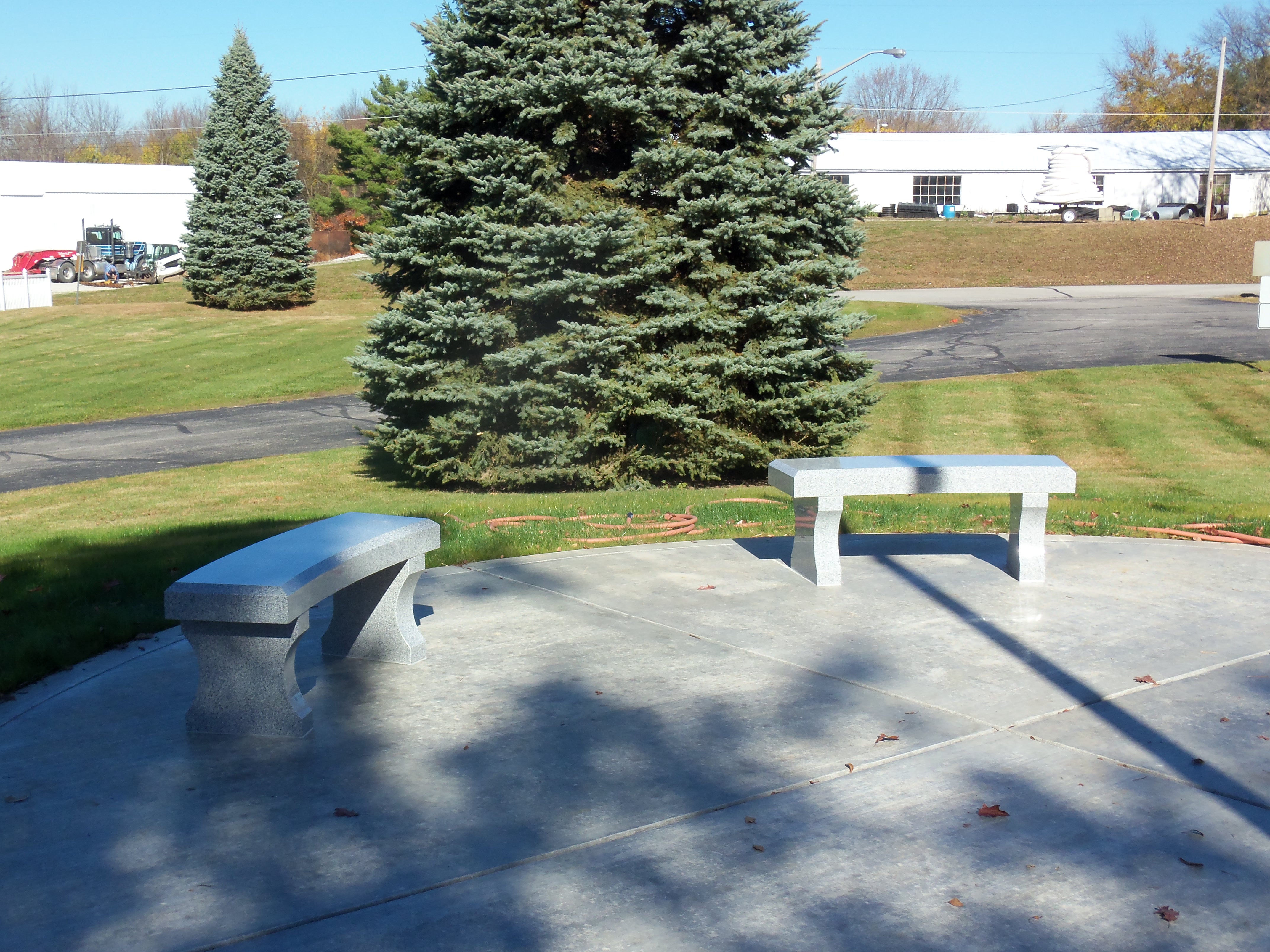 Two grey granite benches with curved legs set on a concrete pad in a park beside a blue spruce