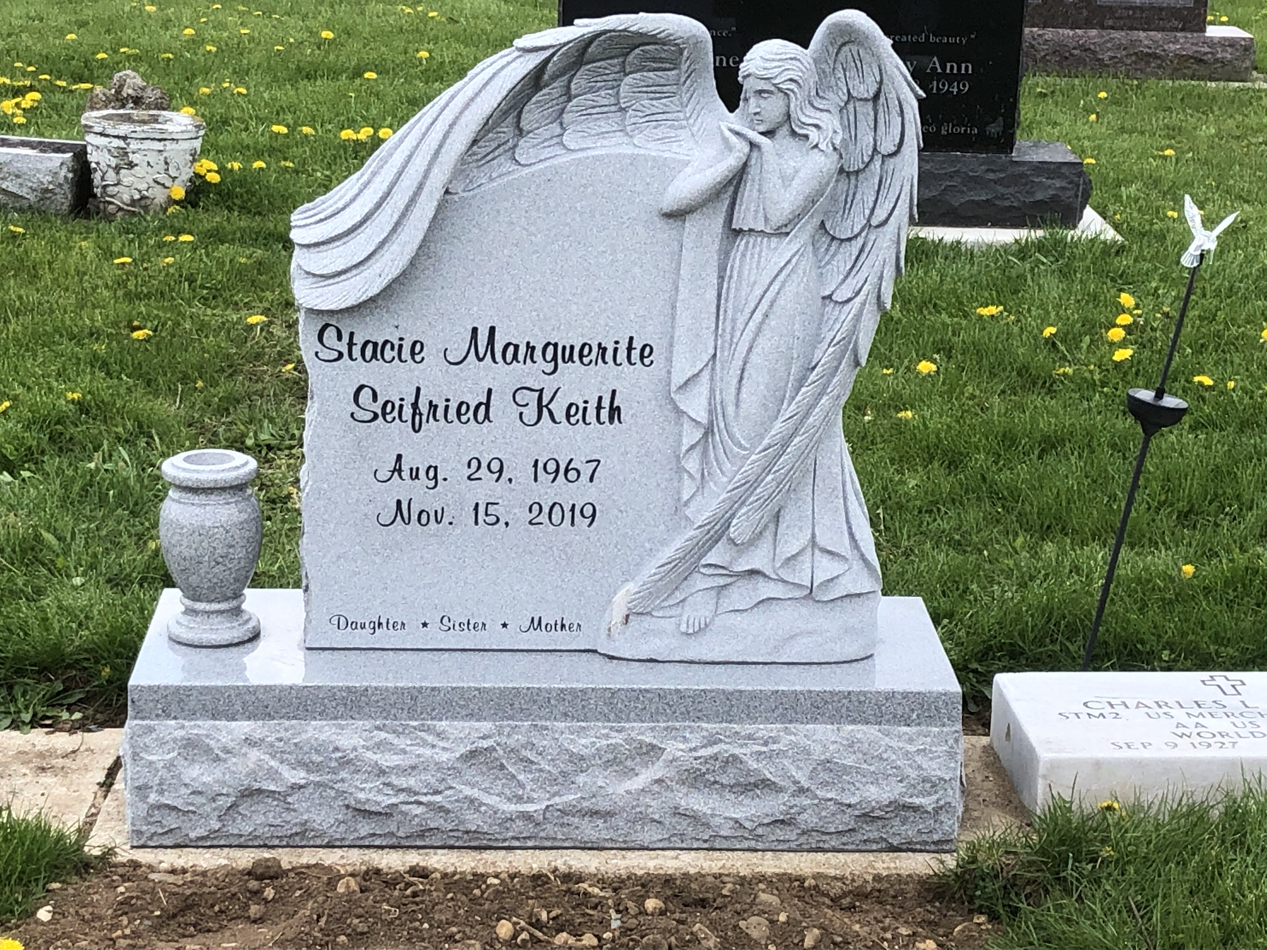 Grey granite upright memorial with an angel carved in deep relief along one side and a draped shoulder, with a matching granite vase